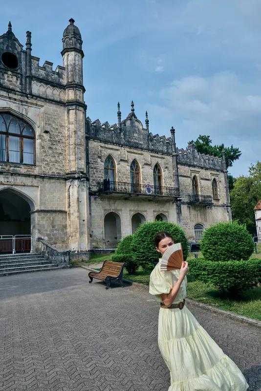 Elegant woman in a pale yellow dress poses in a historic castle courtyard, creating a timeless fashion moment amid stone arches and outdoor manicured garden and warm sunlight.