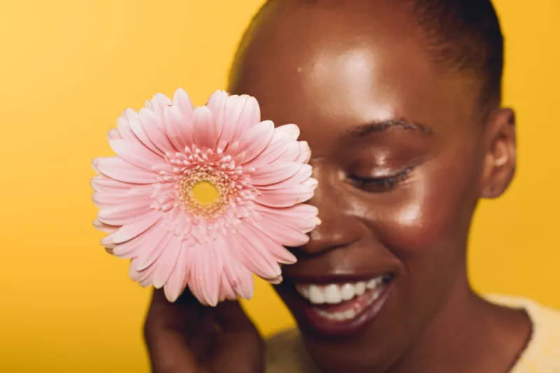 Close-up of a smiling black woman holding a pink flower near her face against a yellow background. Concept of happiness, beauty, floral, and natural skincare for dark skin tone.
