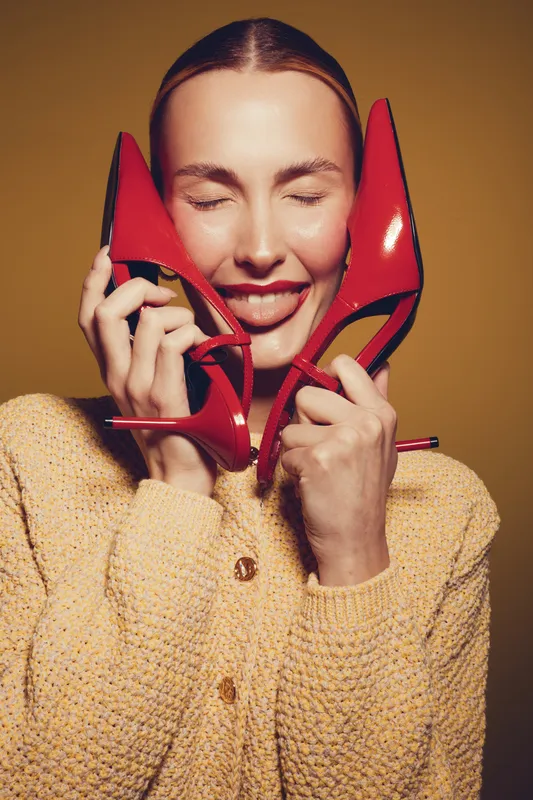 Happy woman with closed eyes holds red high heels near face, smiling with tongue out. Fashion concept with bright shoes and cozy yellow sweater on warm brown background.