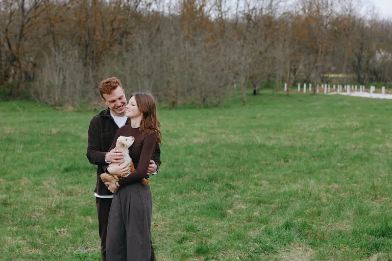 A smiling couple stands in a grassy field, embracing a small dog between them. Soft natural light and a wide rural backdrop convey warmth, companionship, and simple moments of happiness outdoors