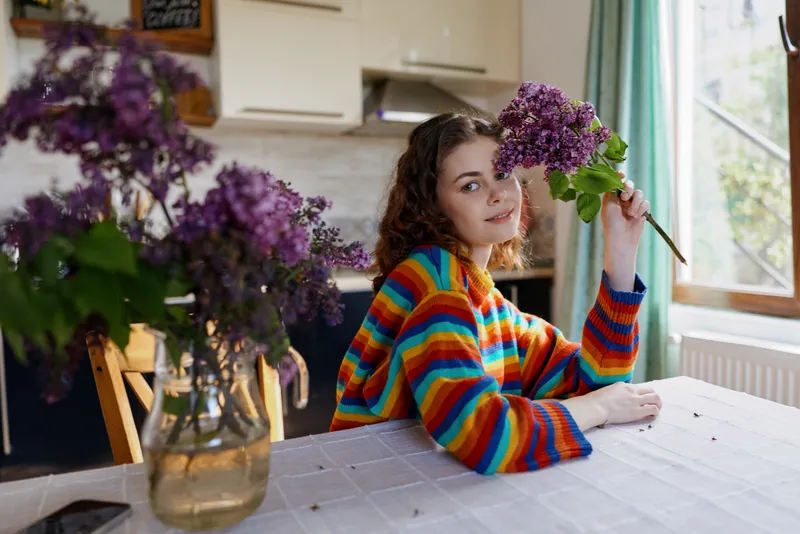Smiling woman with colorful striped sweater sits at a kitchen table, holding a bouquet of lilac flowers. Bright window light creates a cozy home mood, with a simple, everyday elegance.