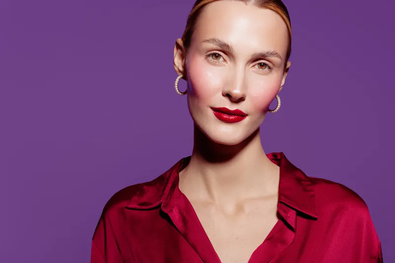 Portrait of confident woman with red lipstick and earrings wearing red satin blouse on purple background. Concept of fashion, beauty, elegance, and modern style in studio lighting.