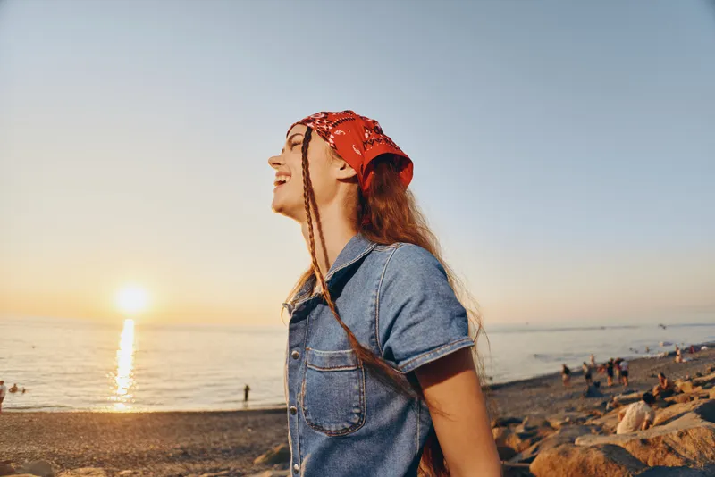 Lifestyle woman with modern boho-western denim street style enjoys sunset by the sea, wearing a red bandana, carefree and relaxed with film color warmth.