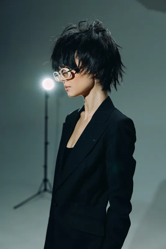 Young woman with black short hair and glasses wearing black suit standing in studio with light background