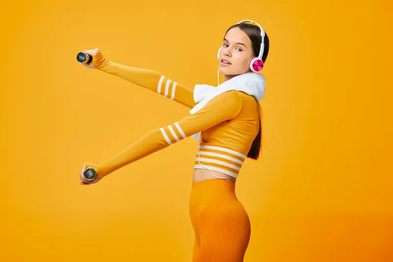 Fitness woman in orange sportwear performs dynamic workout with dumbbells, headphones and a vivid studio backdrop, showcasing active training, cardio focus and athletic energy.