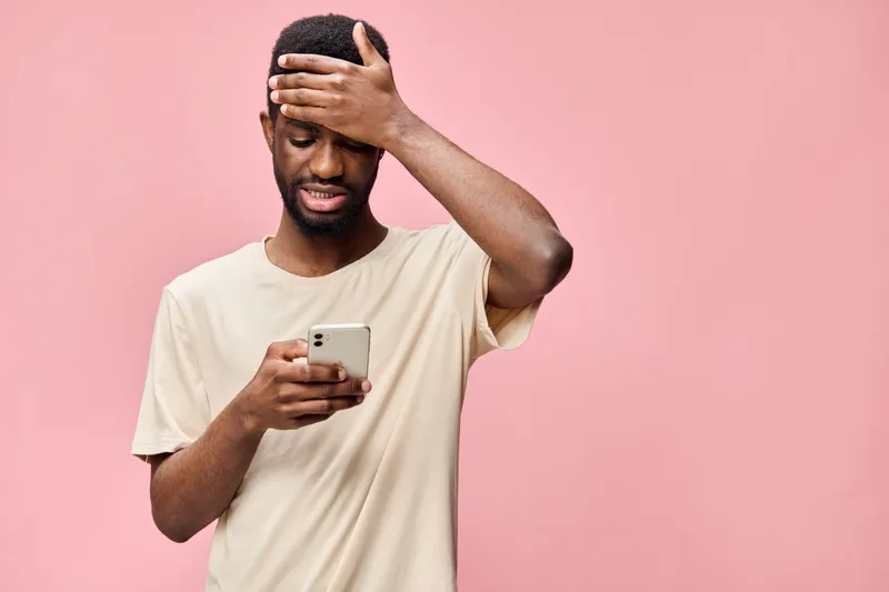 Young African man feeling stressed while reading messages on his smartphone against a pink background, expressing frustration and anxiety in a casual outfit