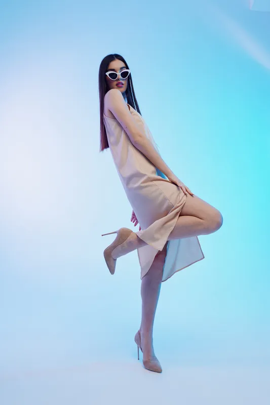 Stylish woman in a beige dress and high heels poses confidently wearing white sunglasses. Studio shot with blue gradient background. Concept of woman beauty fashion elegance.