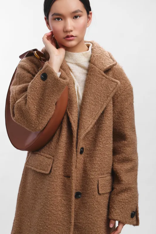 Asian woman wearing a plush tan coat and knit sweater stands in a modern setting, carrying a rounded bag. The image conveys a calm, contemporary fashion portrait with a confident, stylish mood.