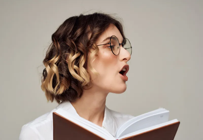 Женщина с вьющимися волосами в очках держит в руках книгу и работает в студии
