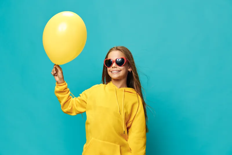 Girl in yellow hoodie holding yellow balloon smiling with sunglasses on blue background for happiness, youth, celebration, party and casual style portrait.