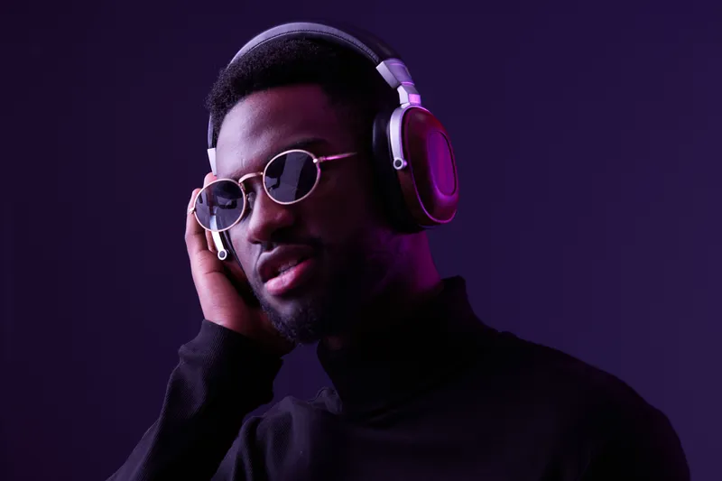 Stylish young Black man wearing headphones and sunglasses, enjoying music against a vibrant purple background, conveying relaxation and modern lifestyle