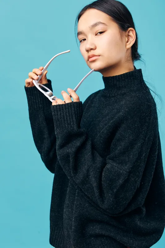 Young woman in black sweater holding eyeglasses with confident look standing against blue background, casual portrait, fashion style and modern beauty concept.