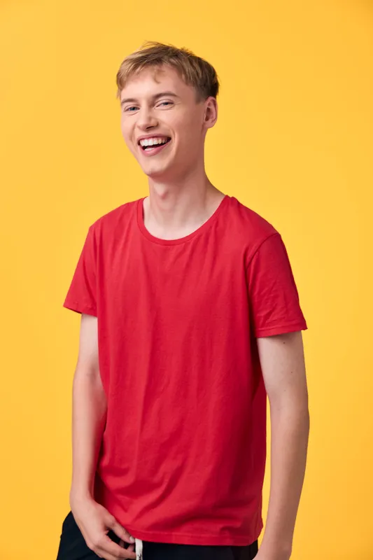 Smiling young man in a red shirt stands against a bright yellow backdrop, conveying casual confidence and approachable style for lifestyle, fashion, or advertising uses.