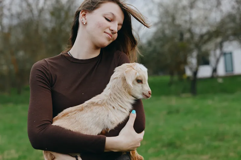 Cheerful woman outdoors, holding a goat kid with affection, gentle hug and sunlight, field and countryside backdrop, calm candid moment, natural colors, warm friendship between human and animal