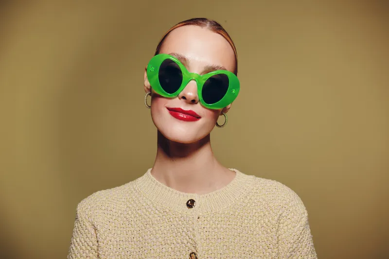 Young woman with slicked back hair wears large green sunglasses and red lipstick. She smiles confidently against a plain beige background. Concept of fashion and style.