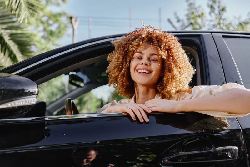 Smiling woman with curly hair leaning out of black car window, enjoying sunny day with vibrant green foliage background and casual beige clothing.