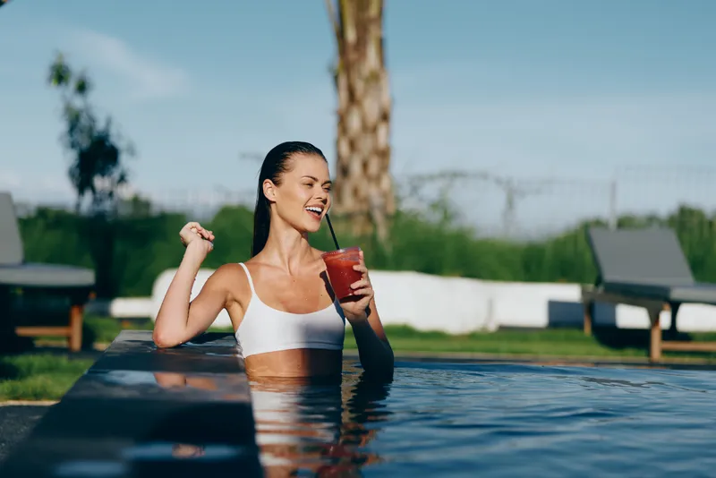 Young woman in white bikini relaxing in outdoor pool drinking red cocktail. Sunny day with clear sky, concept of summer lifestyle, leisure, and vacation time enjoyment.