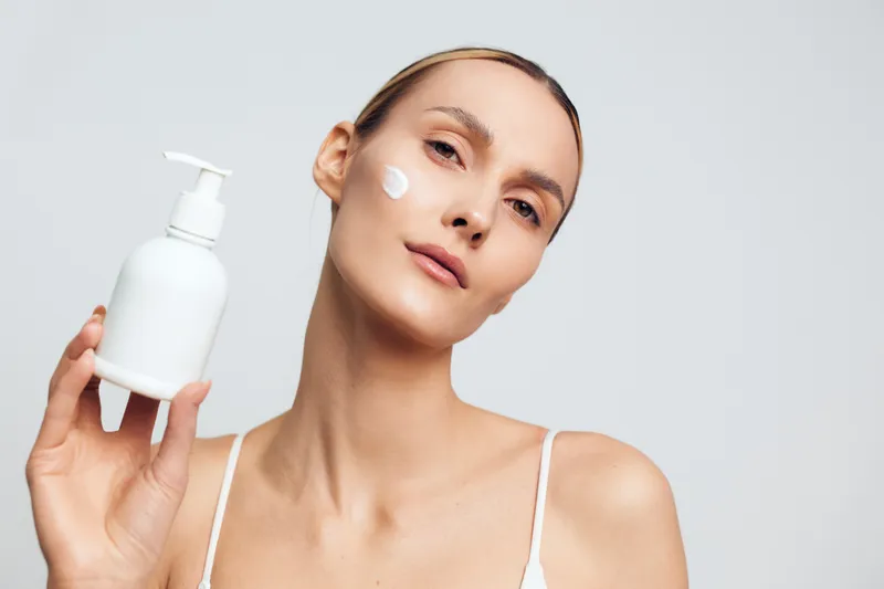 Young woman with clean skin holds white plastic bottle with lotion and applies cream on face isolated on white background with natural expression and minimal makeup.
