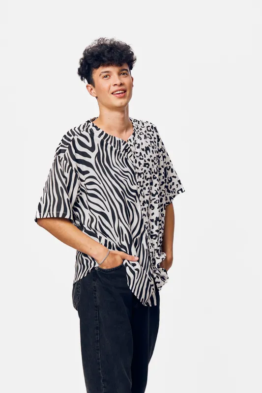 Young man smiling wearing a stylish black and white animal print shirt with casual black pants, standing against a plain white background, modern fashion style.