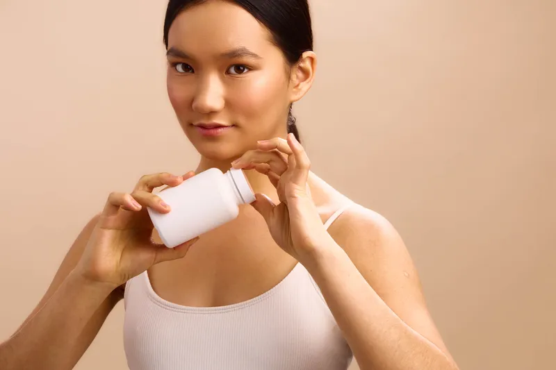 Asian woman pours liquid from a small white bottle into her hand, posing in a calm, focused moment within a neutral studio setting.