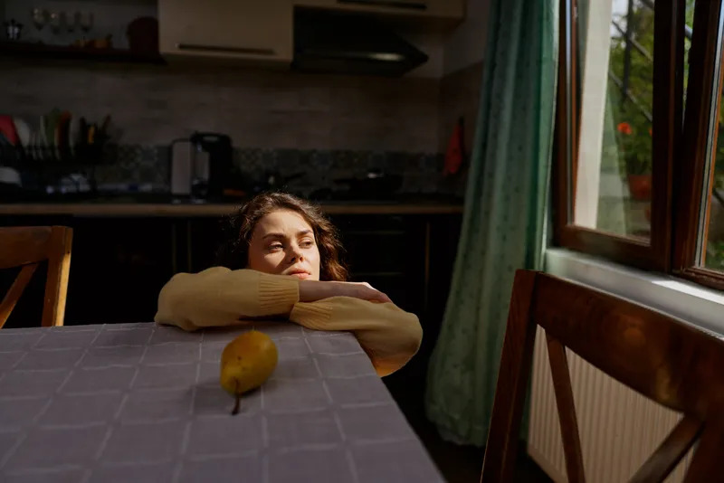Young woman rests her head on her arms across a kitchen table, gazing toward a sunlit window with a single lemon nearby, conveying quiet introspection in a cozy home