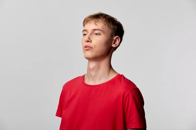 Young man wearing a red t shirt stands against a neutral studio background, thoughtful gaze upward, expressing casual fashion, contemporary youth mood and minimalist simplicity