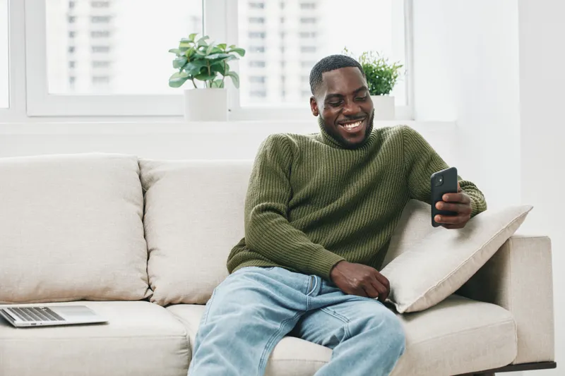 Happy man using smartphone at home, wearing a cozy olive green sweater, comfortable in a bright modern living space, highlighting casual lifestyle and relaxation