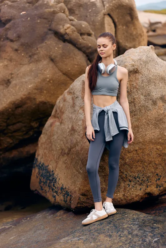 Young woman in activewear standing on rocky coastline, demonstrating fitness lifestyle and healthy living with a confident expression