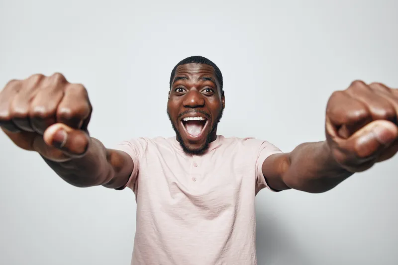 African American man with dark skin wearing light pink shirt showing fists forward with excited expression on white background. Portrait of happy black male with enthusiasm.