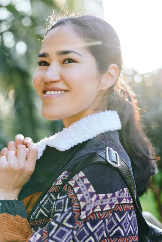 Smiling woman wearing a patterned sweater and backpack outdoors with sunlight, greenery and a warm, approachable mood, showcasing casual fashion and travel style for lifestyle stock
