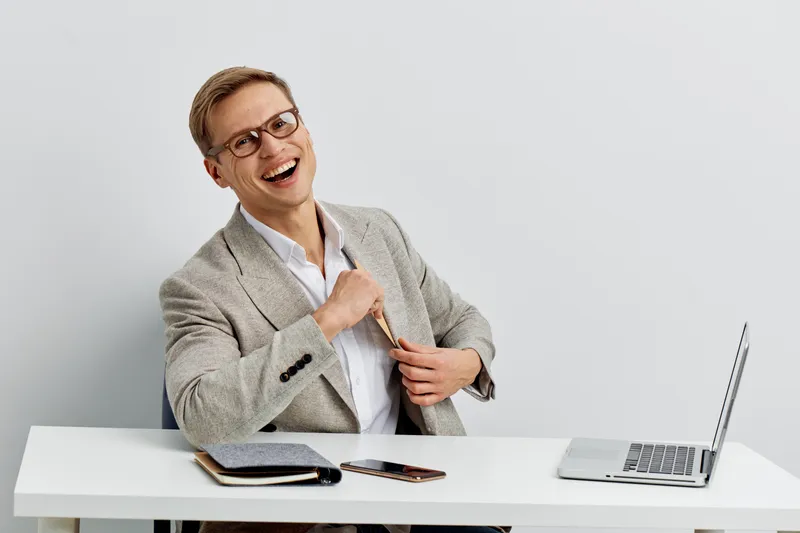 Confident middle-aged woman in business attire smiling cheerfully at camera while sitting at a white desk with a laptop and smartphone, isolated on plain light gray background, studio portrait. People
