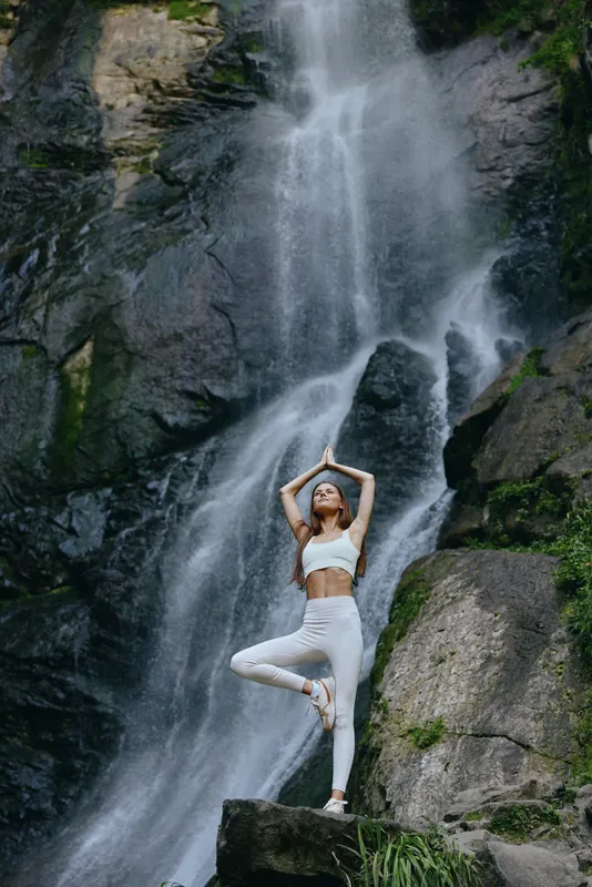 Young woman practicing yoga on a rock near a flowing waterfall surrounded by lush green nature in a serene outdoor setting on a bright day.