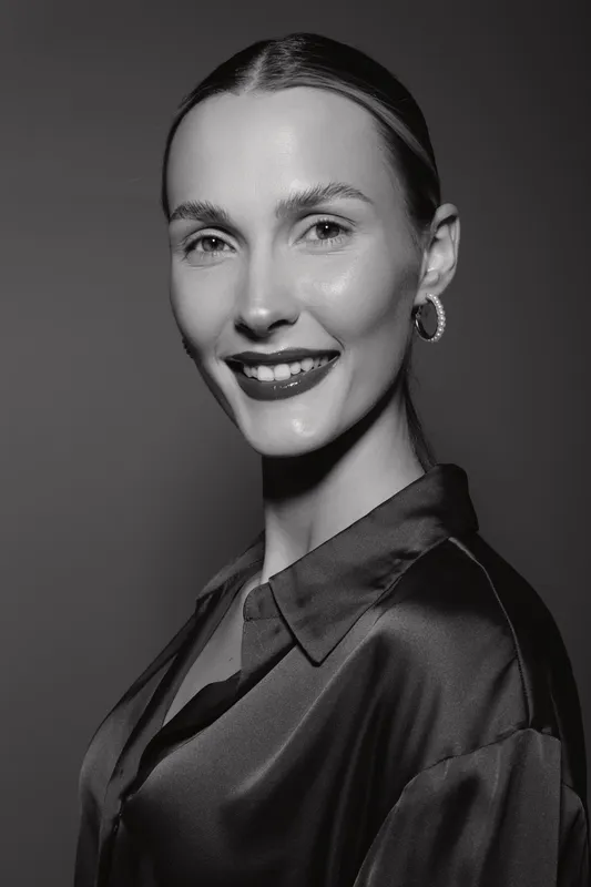 Young white woman with fair skin and slicked back hair smiles confidently wearing hoop earrings and satin blouse. Studio portrait in black and white with soft lighting and elegant mood.