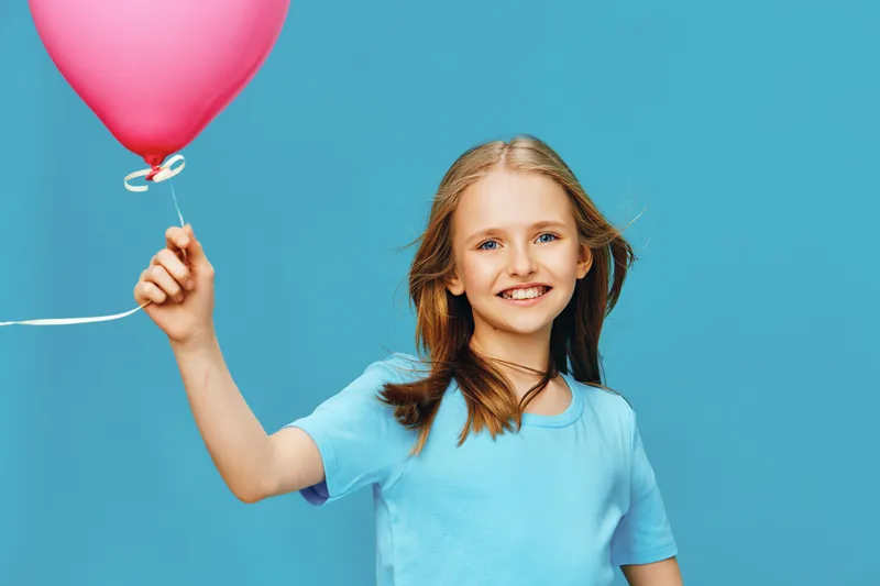 Happy girl with long hair holding a pink balloon against a bright blue background, wearing a light blue t shirt, expressing joy and excitement