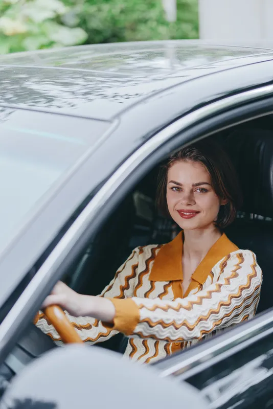 A confident woman driver sits behind the steering wheel in a car, smiling warmly as she prepares for a journey, radiating readiness, focus, and joyful anticipation of travel.