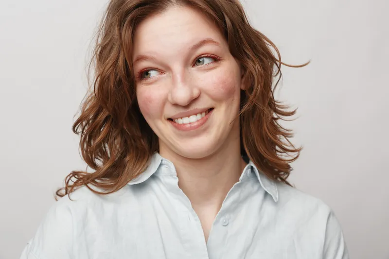Smiling young woman with wavy brown hair wearing a white shirt looks to the side with a playful expression. Studio portrait on a light grey background. Concept of happiness and casual style.