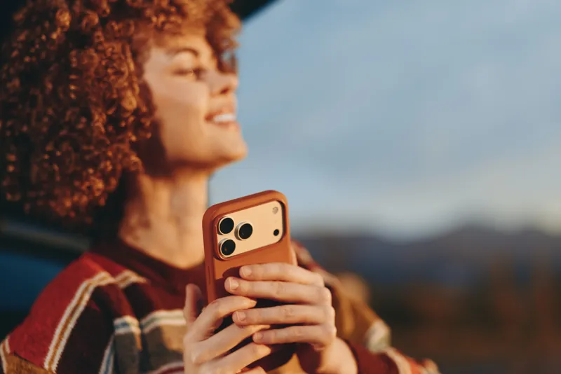 Woman lifestyle outdoors, portrait of curly haired woman holding smartphone, warm sweater, looking away using phone, golden sunset light, mountain background, relaxed mood in fresh air