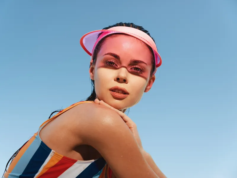 Close up portrait of a young woman in a colorful striped swimsuit under bright sun, wearing a pink visor, sunlit skin, confident gaze, and carefree summer vibe.