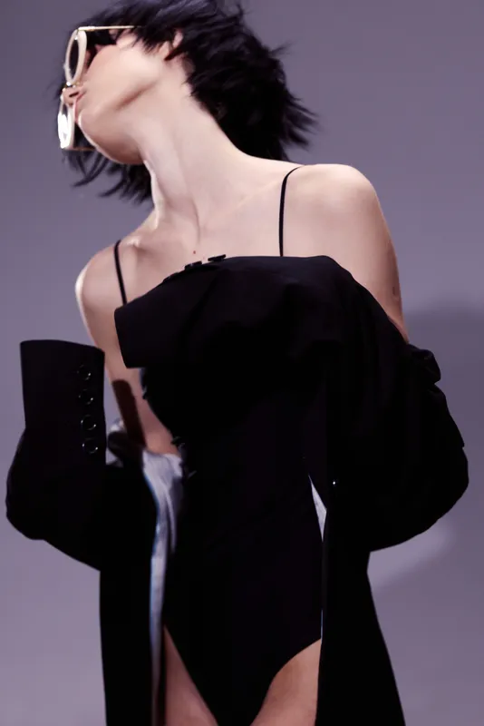 Stylish woman wearing black dress and oversized jacket in dynamic pose with sunglasses, modern fashion concept