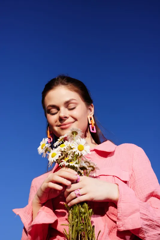 Young woman with flowers in vibrant pink outfit against a clear blue sky, expressing joy and tranquility, showcasing beauty in nature and seasonal fashion