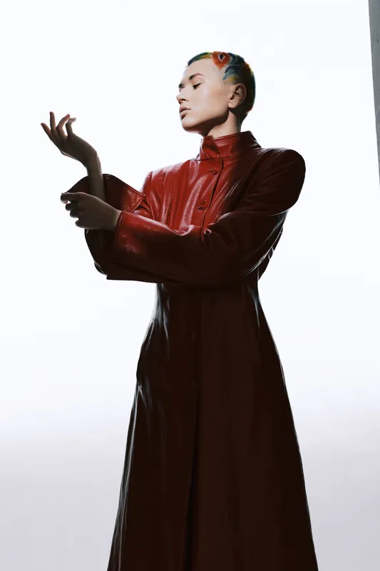 Woman with colorful short hair wearing a long red leather coat posing gracefully against a bright white background, showcasing modern fashion and style details.