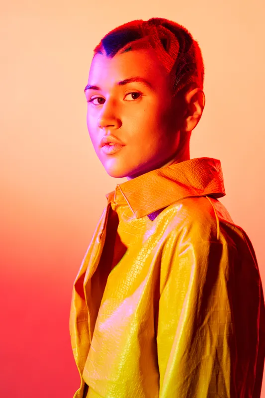 young woman with braided hair and shiny yellow jacket looks confidently with orange and pink gradient lighting highlighting her face and outfit in a modern studio setting
