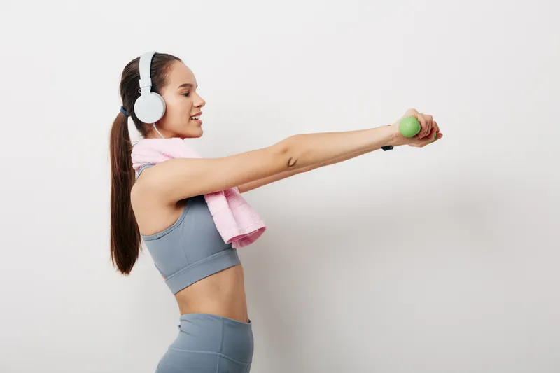 Young woman fitness workout in sportswear holding small green dumbbell, wearing headphones and pink towel, performing front raise exercise against plain white background.