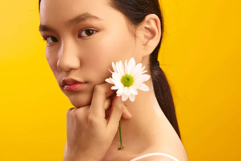 Asian woman stands against a vibrant backdrop with a flower, presenting a calm and graceful portrait that highlights natural beauty and a creative, upbeat mood.