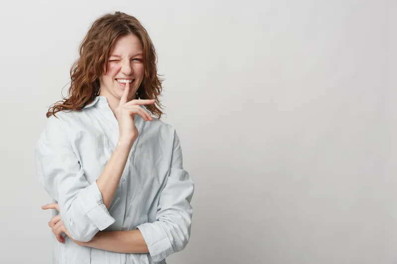 Young woman with curly hair in white shirt makes shushing gesture with finger on lips, eyes closed, isolated on light background. Concept of silence, secrecy, and quiet communication.