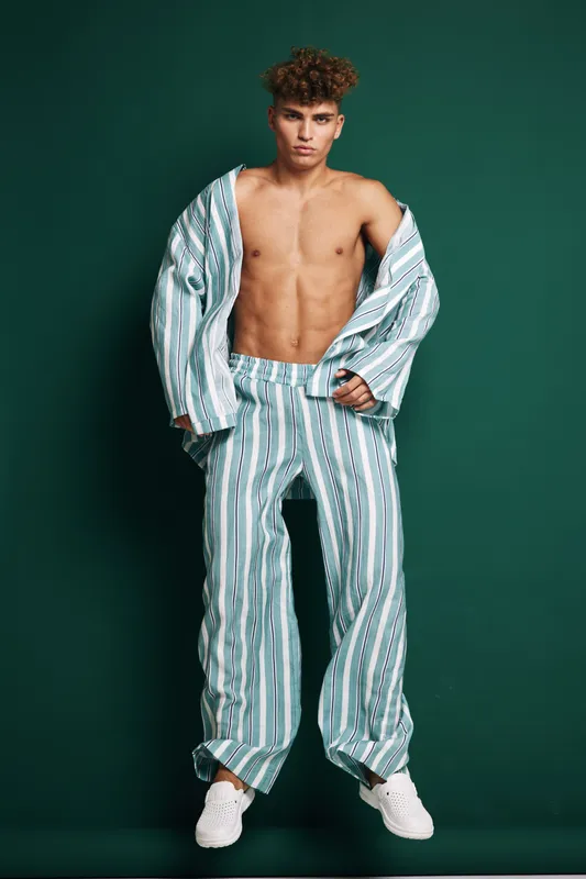 Young man standing against green background wearing striped open shirt and matching trousers with white sneakers barefoot relaxed confident casual look stylish fashion model pose