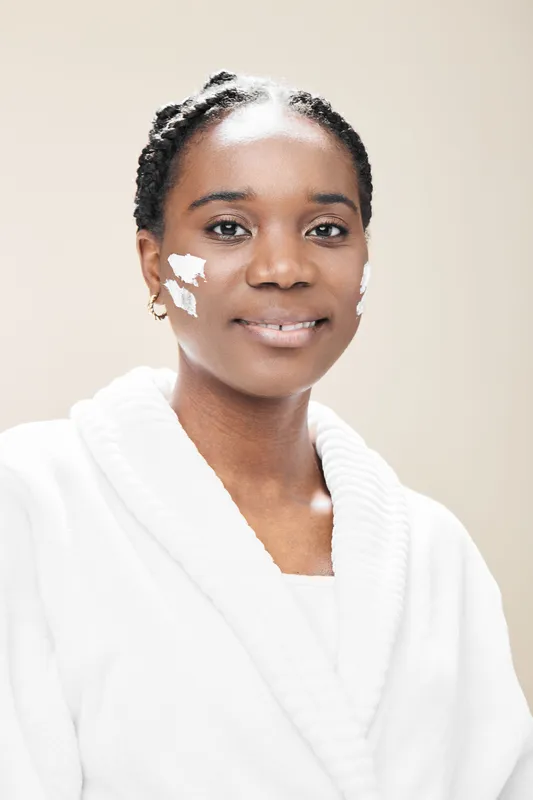 Young woman with dark skin smiling while applying face cream, wearing a white robe, with a neutral background, representing skincare and self care rituals
