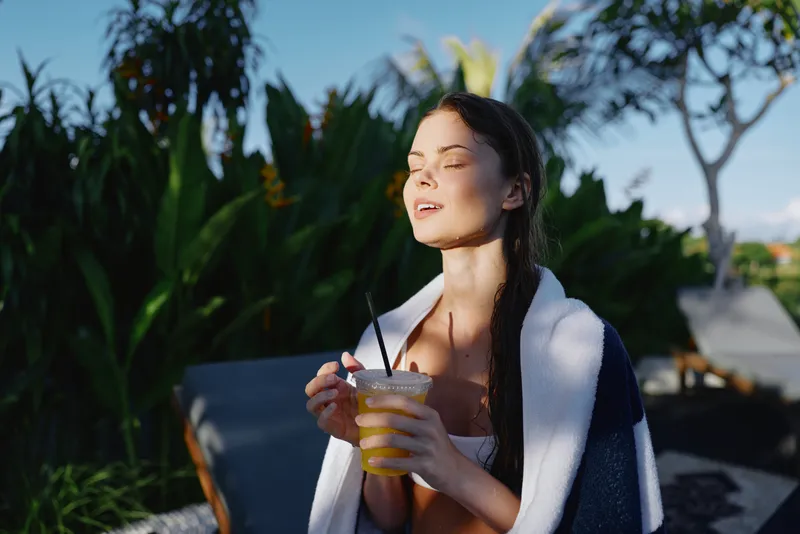 A relaxed woman enjoys a cold beverage by a sunny outdoor pool, wrapped in a towel after a swim. The scene conveys leisure, wellness, and calm in a bright, vacation mood.