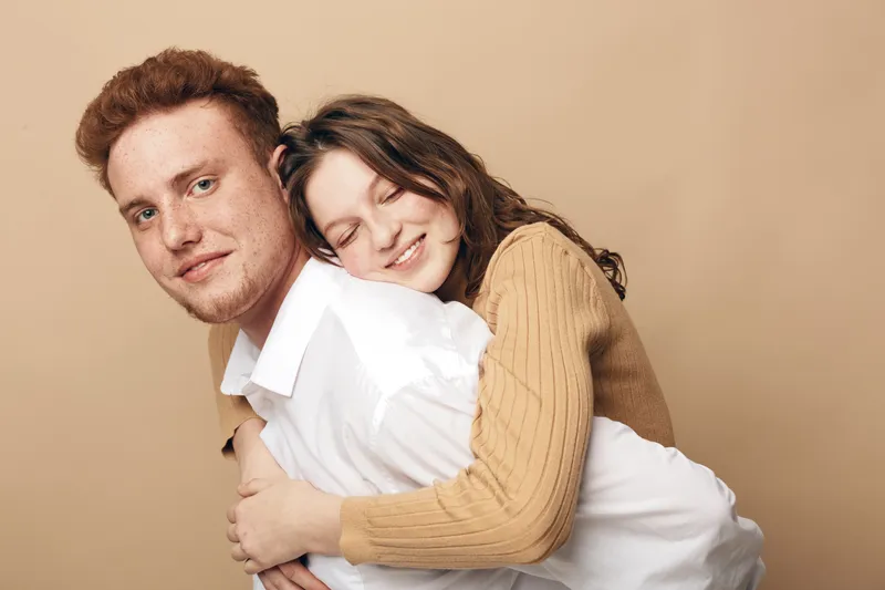 Smiling young woman in beige sweater hugs happy man in white shirt from behind on beige background. Concept of love, friendship, affection, and close relationship between couple or friends.