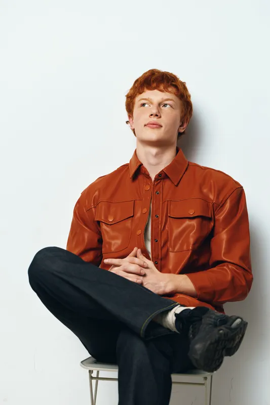 Young man with red hair wearing brown leather jacket and black pants sitting on stool with legs crossed looking sideways on white background.